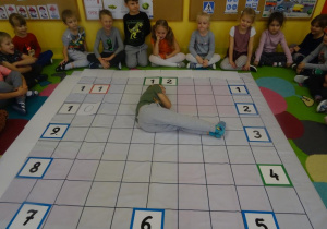 Grupa dzieci siedzi wokół rozłożonej na dywanie macie do kodowania. Na macie ułożony jest zegar z tabliczek. Chłopiec leży na macie ułożony w literkę L wskazując tym samym godzinę 3.